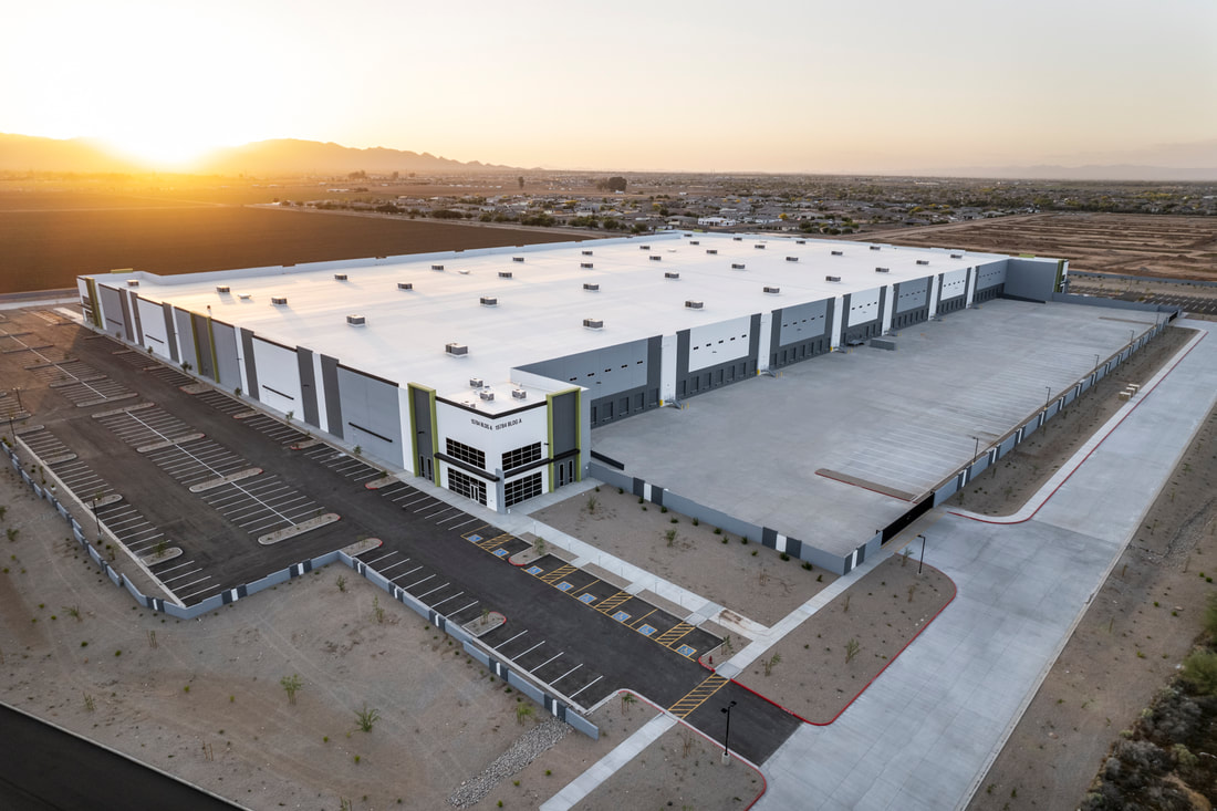 Aerial view of a large modern industrial building with a white roof, multiple loading bays, and surrounding parking areas, set against open fields and a sunrise over distant mountains.