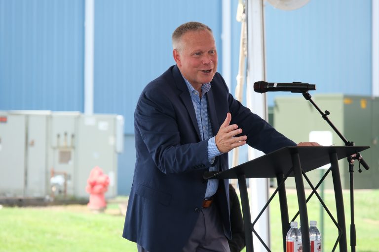 CEO TIm Mills delivering a speech at a podium, celebrating the 9 millionth transformer milestone in a decorated factory setting.