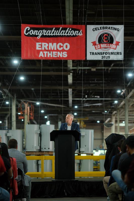 A photo of Tim Mills standing behind a podium on stage address a ground in front of him. Tim is centered in the photograph with a celebration banner above it his and electrical transformers serving as a backdrop.