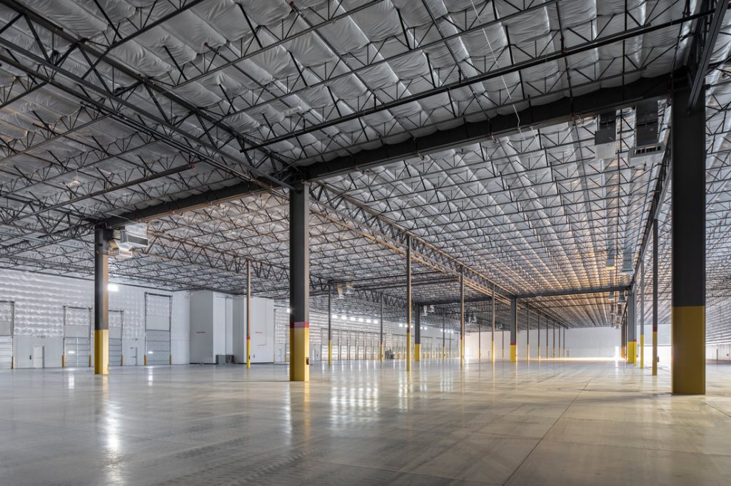 Interior view of a large, empty industrial warehouse with high ceilings, exposed steel trusses, polished concrete floors, and multiple loading bay doors along the far wall.