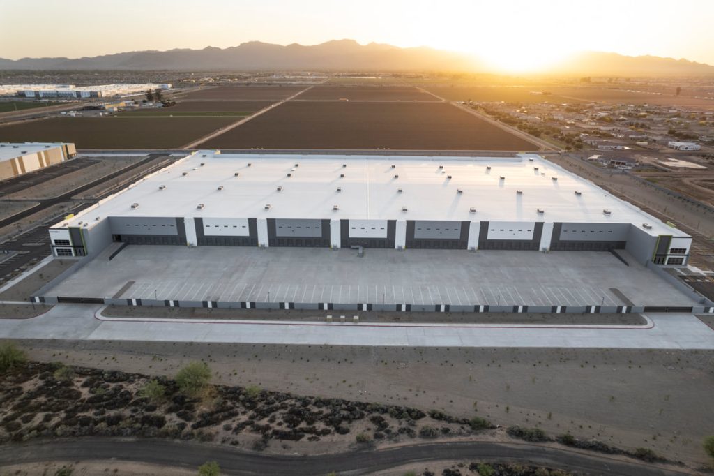 Aerial view of a large, modern industrial building with a white roof and multiple loading bays, surrounded by open land and agricultural fields, with mountains and a sunrise in the background.