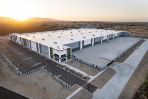Aerial view of a large modern industrial building with a white roof, multiple loading bays, and surrounding parking areas, set against open fields and a sunrise over distant mountains.